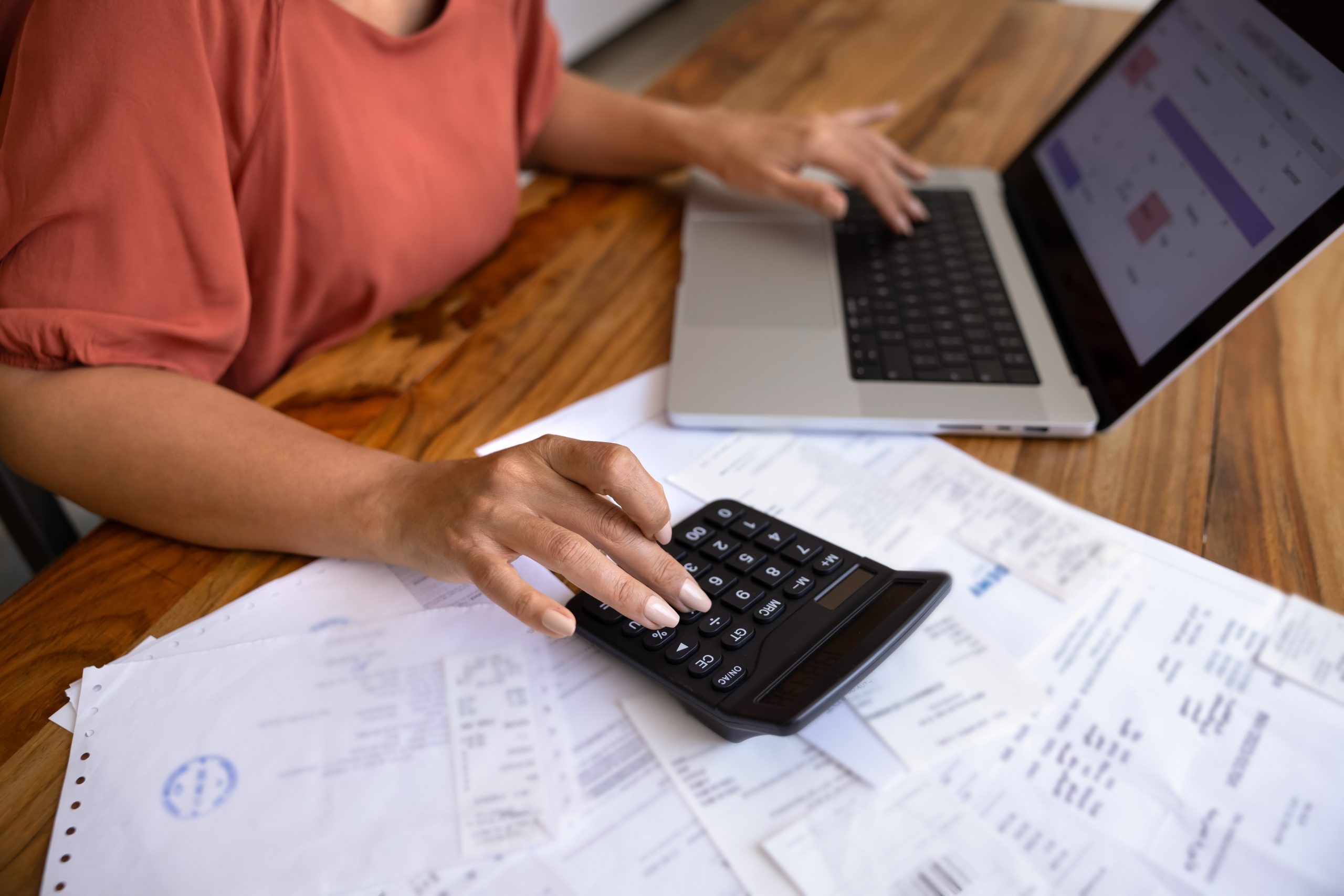 Foto recortada de uma contadora digitando em uma calculadora e um laptop em uma mesa de trabalho com uma pilha de faturas, comprovantes de pagamento, recibos e documentos financeiros em papel, controlando as despesas. 